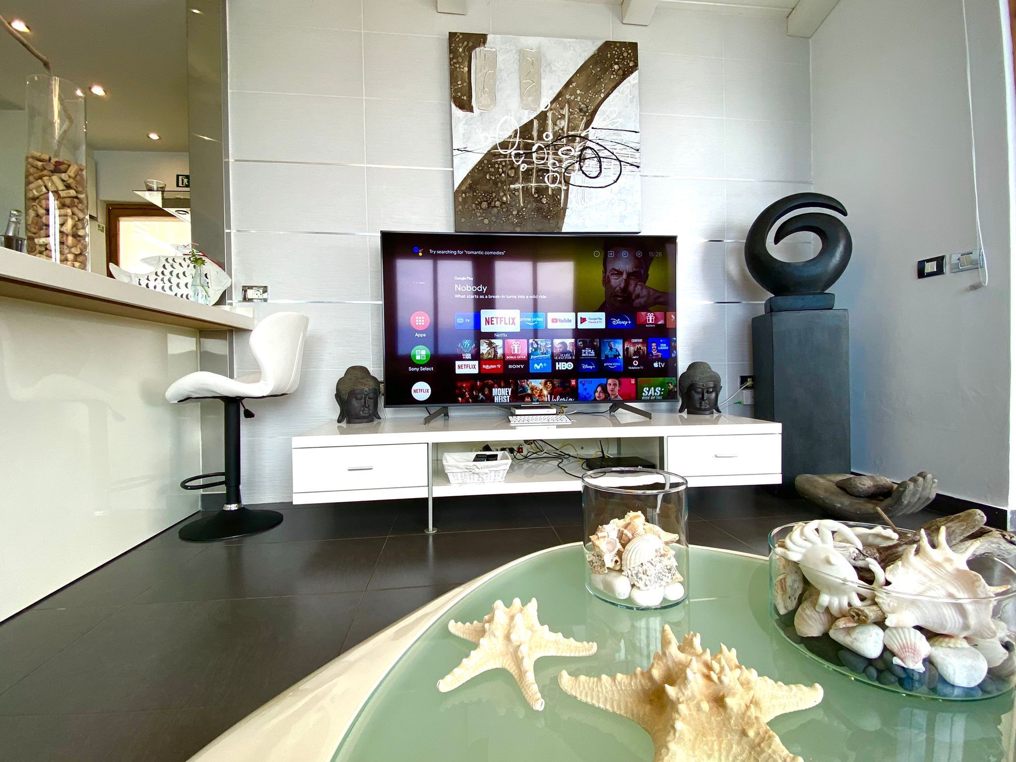 A modern living room with a flat-screen TV displaying a streaming service interface, a white sofa, a glass coffee table with seashells, and a minimalist decor.
