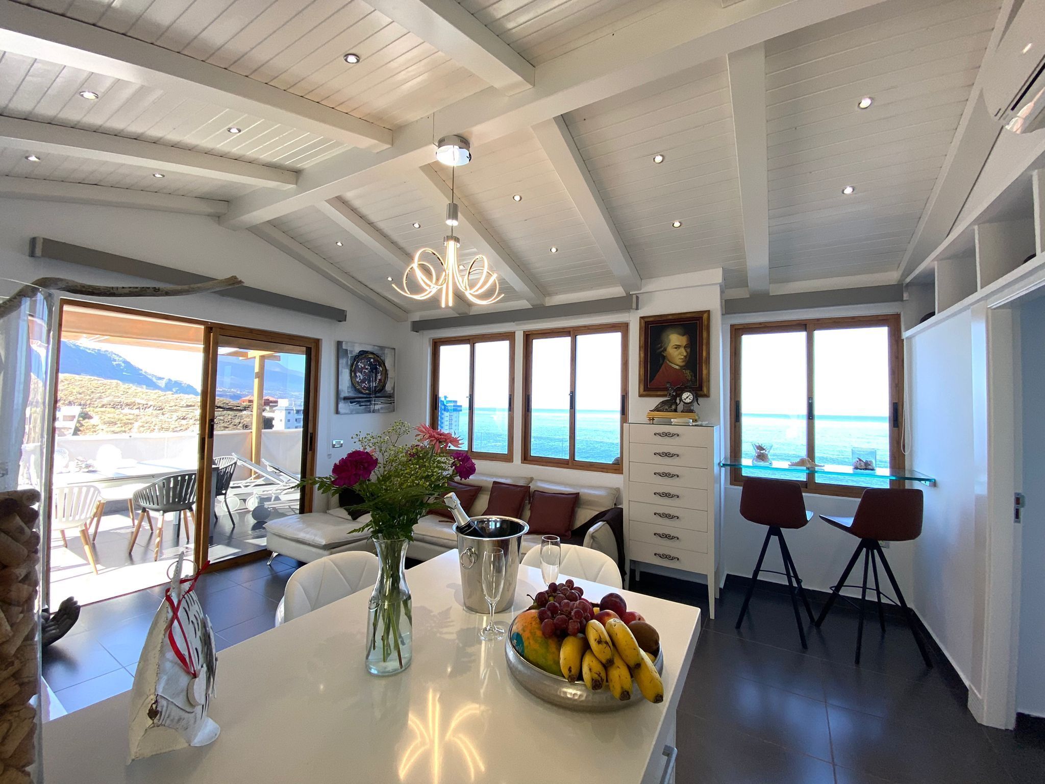 A modern dining area with a view of the sea, a chandelier, and a dining table set with fruits and a champagne bottle.