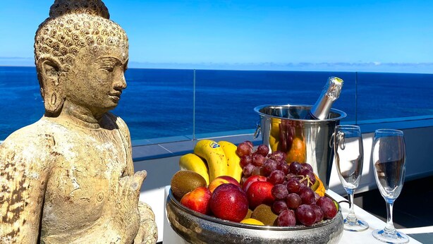 Een Boeddhabeeld, een schaal met fruit, een champagnefles en glazen op een tafel met uitzicht op de oceaan.