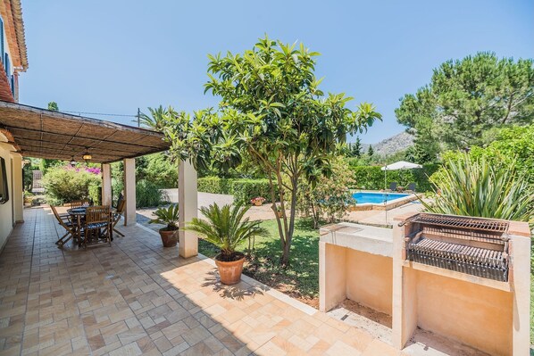 Un patio con mesa y sillas, una parrilla, una piscina y abundante vegetación.