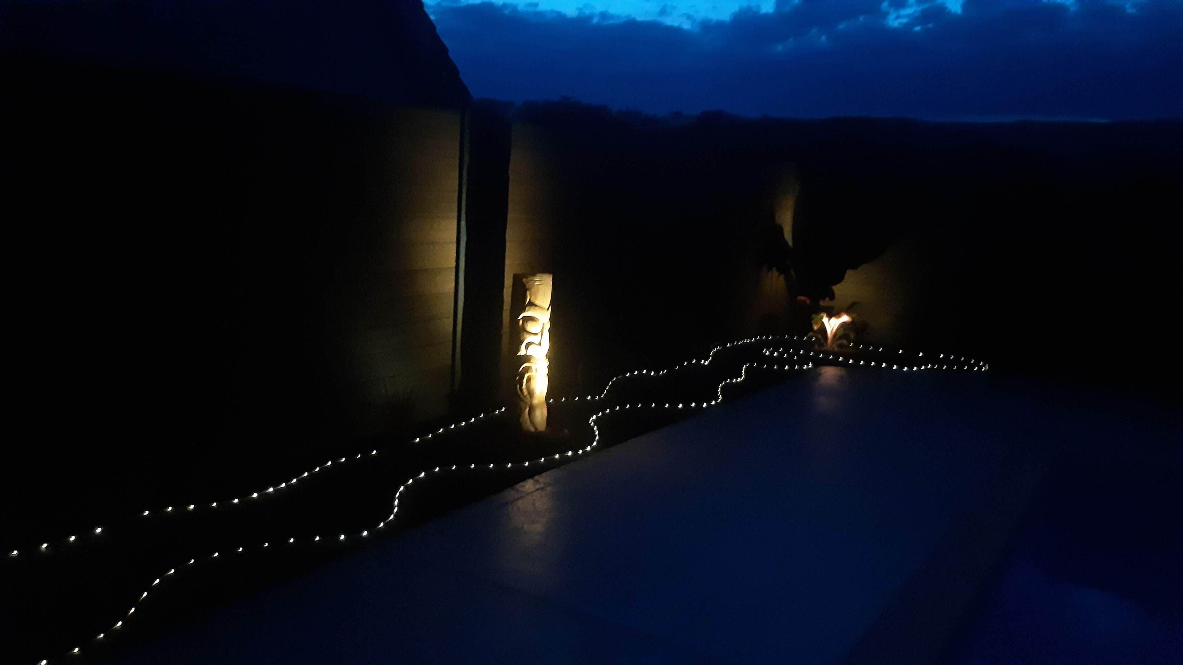 Un chemin faiblement éclairé, bordé de guirlandes lumineuses, qui mène à l’entrée d’un bâtiment.