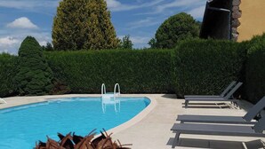 A swimming pool with a clear blue water, surrounded by a hedge and trees. There are lounge chairs and a small table with a plant.