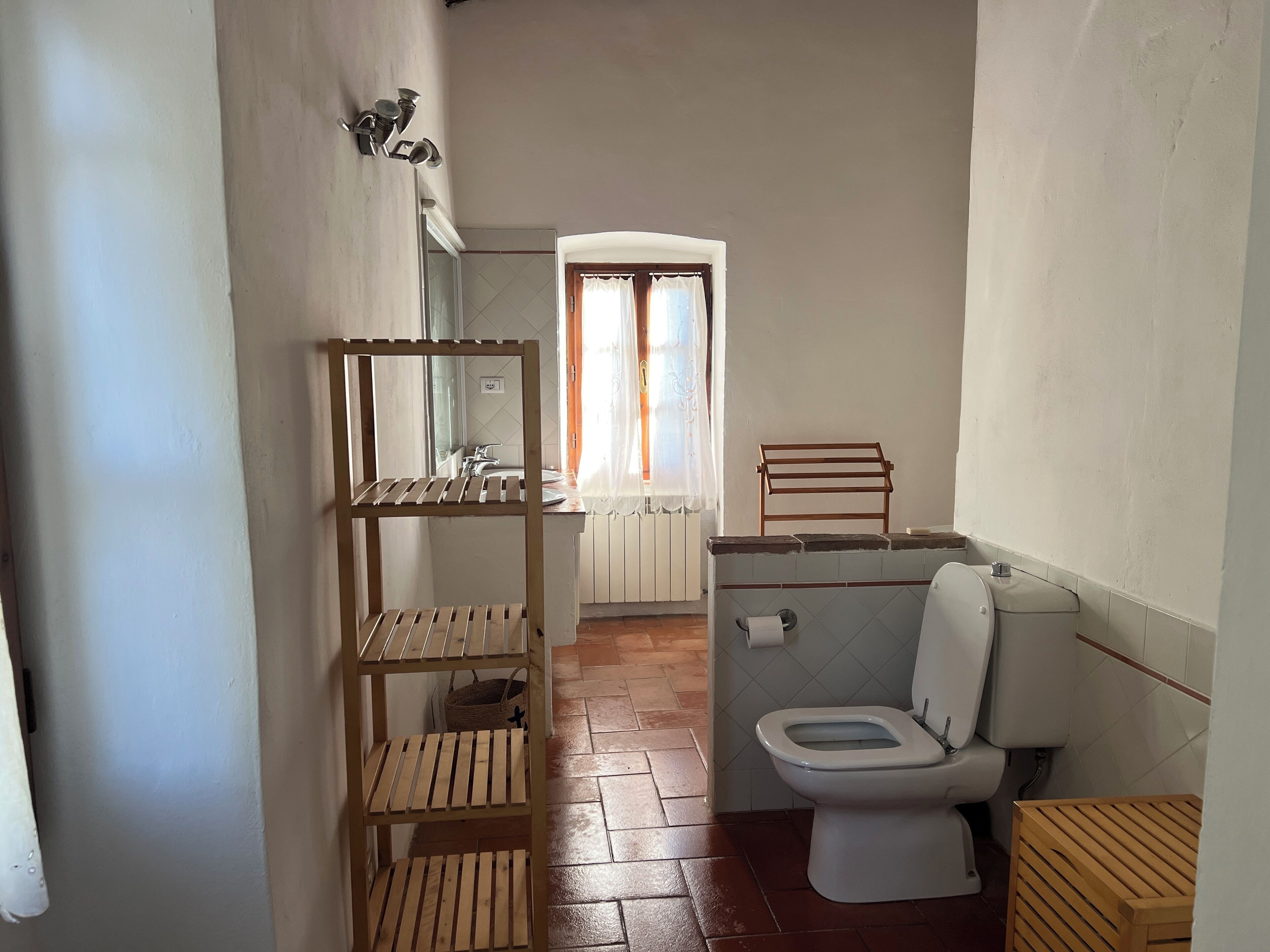 A small bathroom with a wooden shelf, a toilet, and a window with curtains.