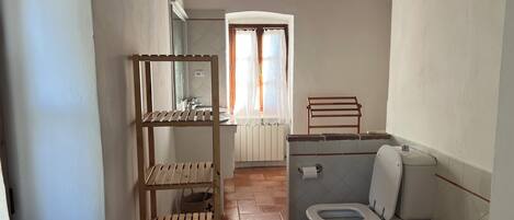 A small bathroom with a wooden shelf, a toilet, and a window with curtains.
