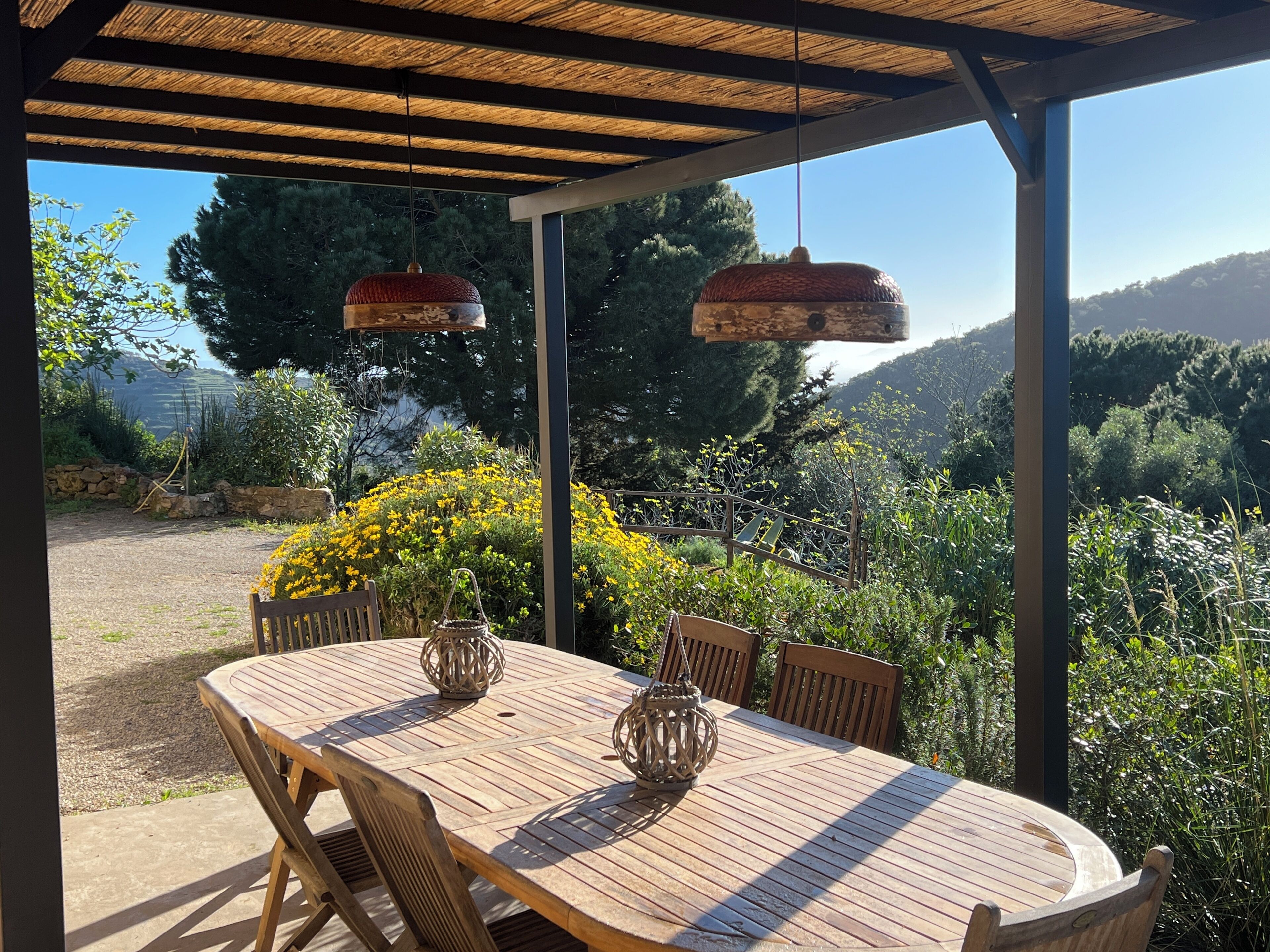 Een houten buitentafel met stoelen en hangende lantaarns onder een pergola, omringd door groen en uitzicht op heuvels.