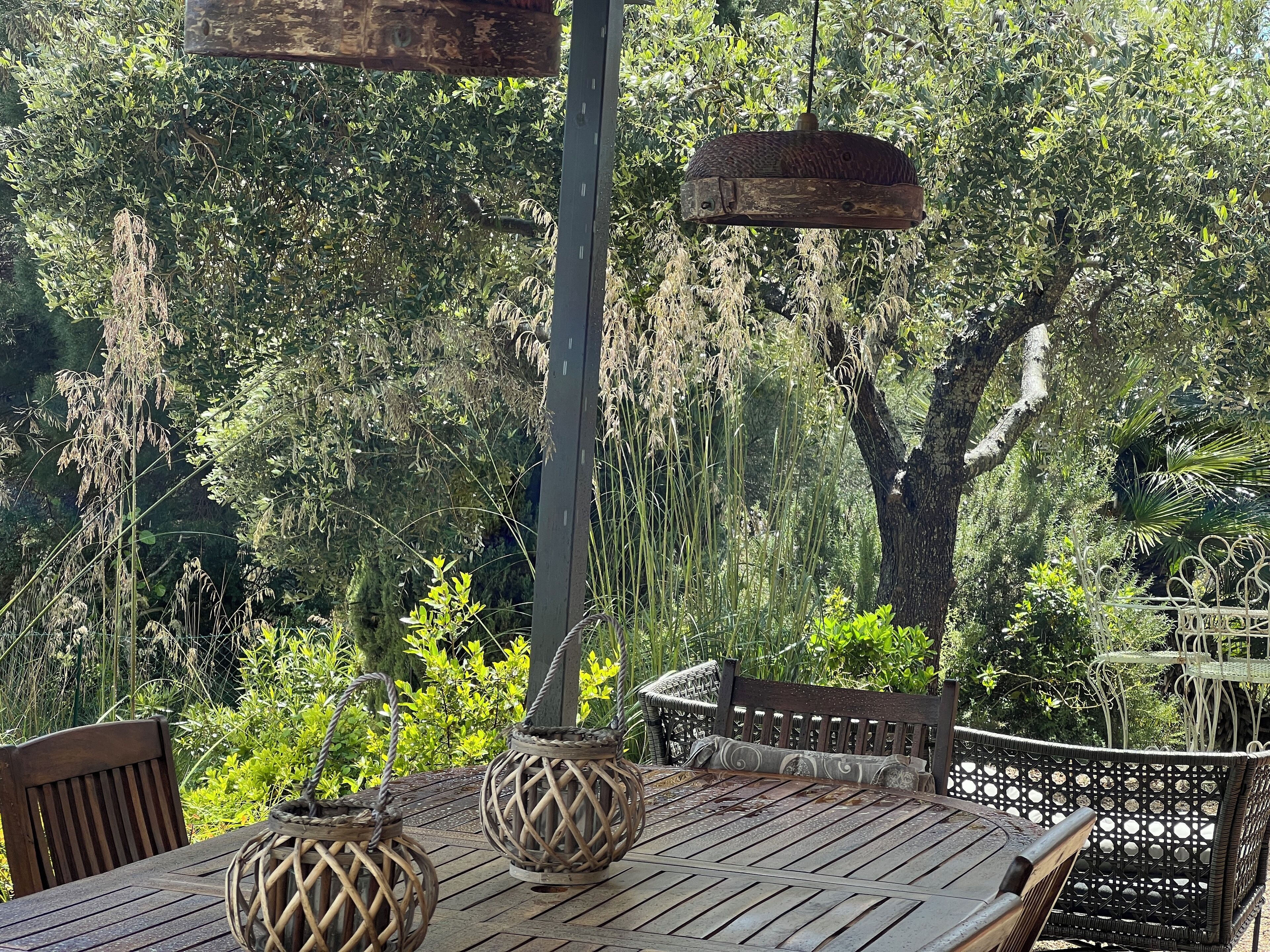 Une table en bois, des chaises en osier et deux lanternes tressées, le tout placé devant un décor de feuillage dense et agrémenté d’un luminaire suspendu.