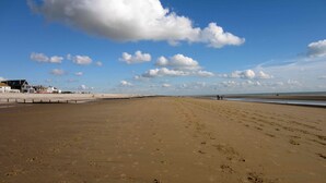 Una amplia playa de arena con huellas, cielo azul despejado y algunos edificios al fondo.