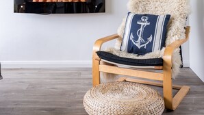 Una silla de madera con un cojín mullido y otro cojín con diseño de ancla, un otoman tejido, un televisor de pantalla plana montado en la pared, y un letrero que dice "CAMBER SANDS".