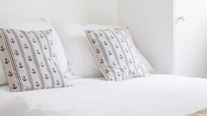 Una cama bien hecha con ropa de cama blanca y almohadas estampadas, una decoración de ancla de madera en la pared y un armario blanco con puerta.