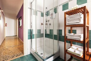 Ein Hotelbad mit einer gläsernen Duschkabine, einem hölzernen Handtuchhalter mit gefalteten Handtüchern und einem gefliesten Boden mit Grün-Weiß-Muster.