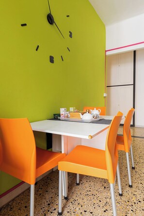 Ein Essbereich mit einem weißen Tisch und orangefarbenen Stühlen, einer Uhr an der Wand und einem Tablett mit Teekanne und Tassen.