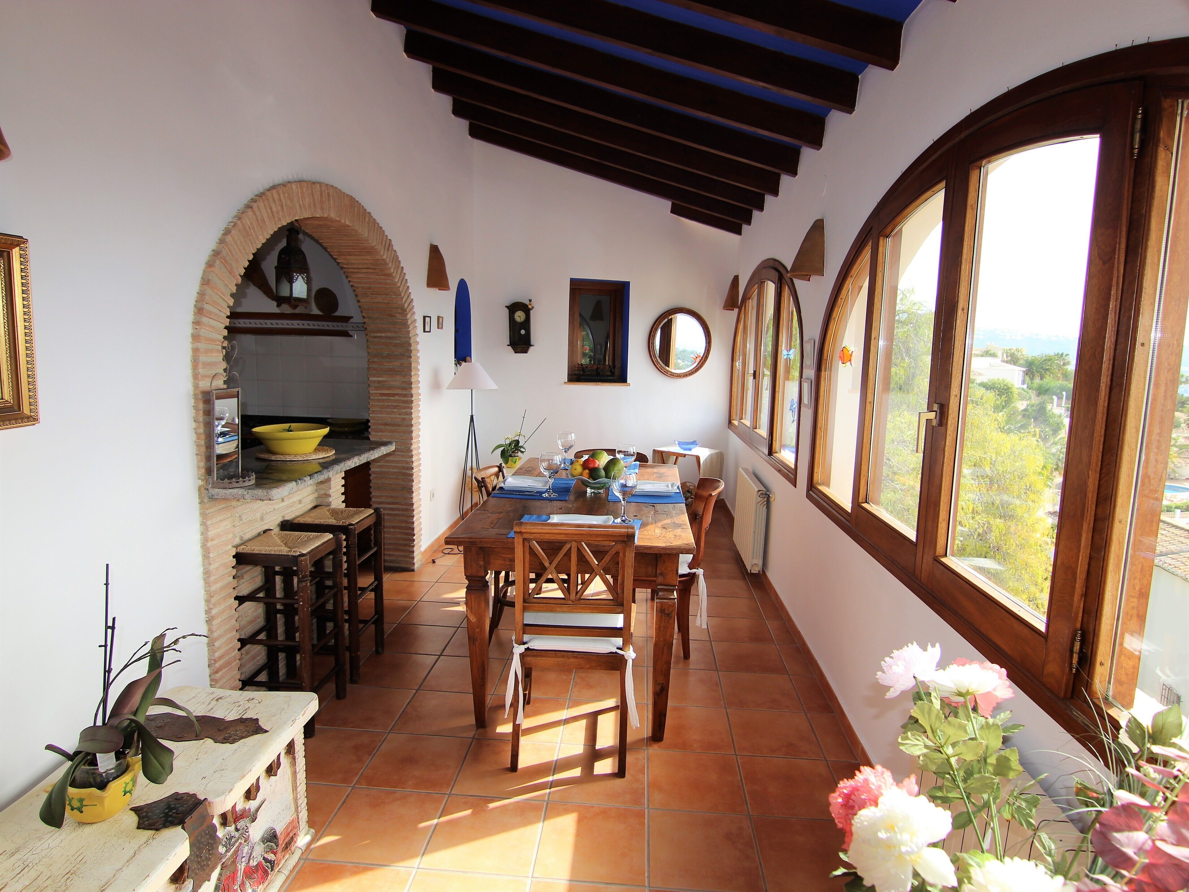 A dining area with a wooden table and chairs, a kitchen with a yellow bowl, and large windows with a view.