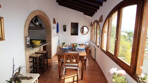 A dining area with a wooden table and chairs, a kitchen with a yellow bowl, and large windows with a view. - House / Villa - MORAIRA (Moraira)