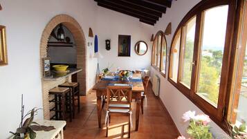 A dining area with a wooden table and chairs, a kitchen with a yellow bowl, and large windows with a view.