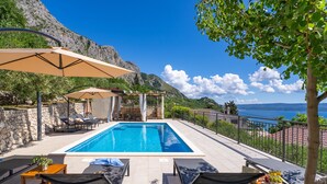 Un espace aménagé au bord de la piscine, avec des chaises longues, une table en bois agrémentée d’une plante, et une vue sur la mer et les montagnes.