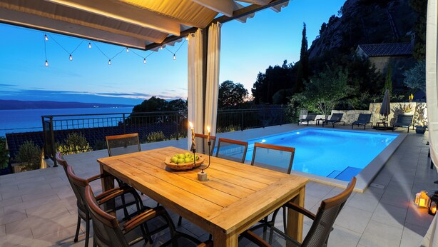 Une table en bois, entourée de chaises, et un saladier rempli de fruits, le tout placé de manière à offrir une vue sur une piscine, la mer et les montagnes.