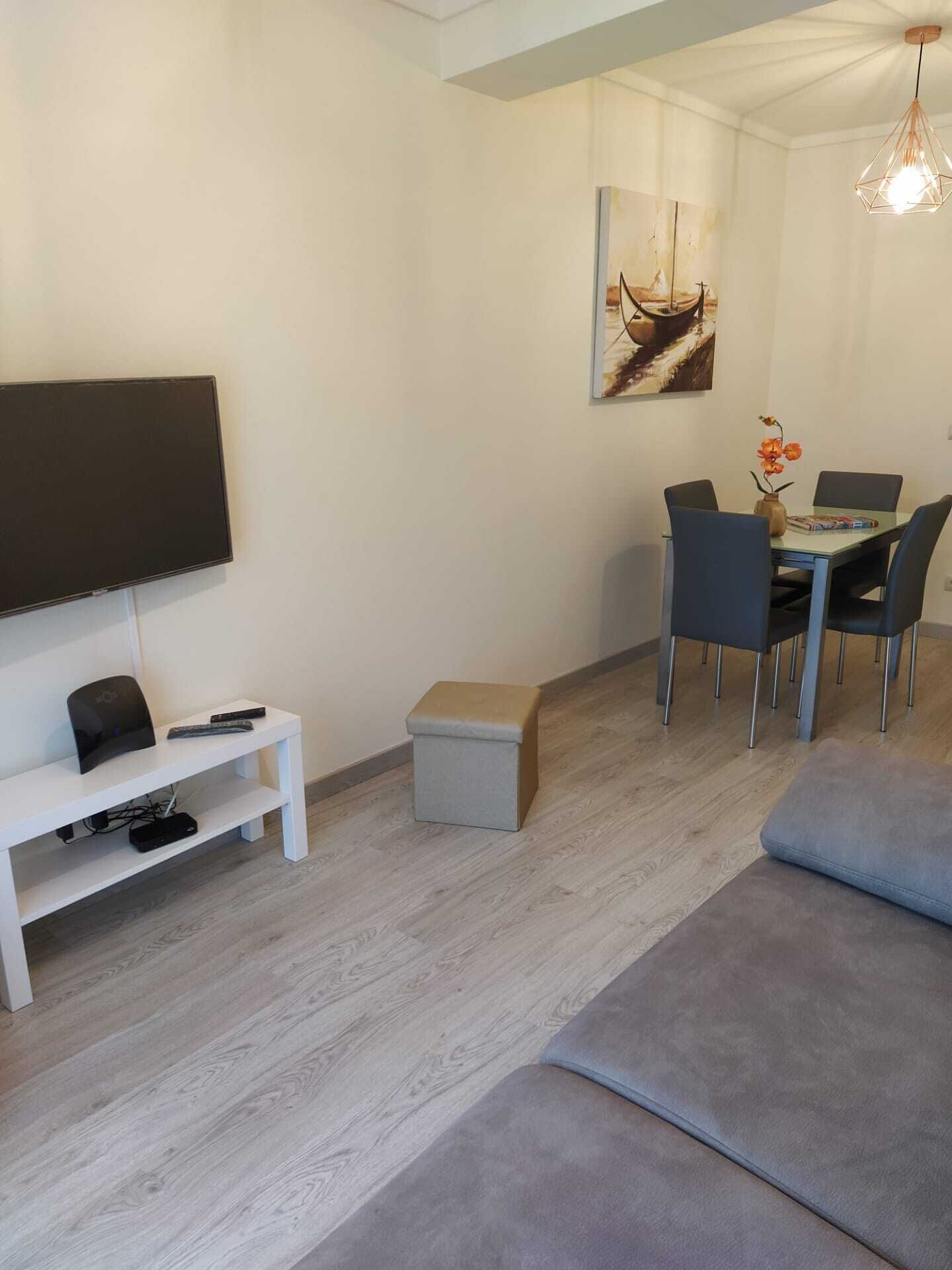 Een moderne woonkamer met een flatscreen televisie, een eettafel met stoelen, een klein voetenbankje en een bank.