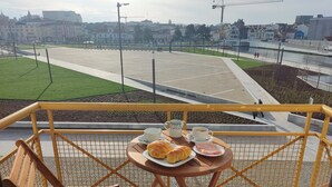 Un balcón con una mesa dispuesta para el desayuno, incluyendo croissants y café, con vistas a un paisaje urbano con un río y edificios.