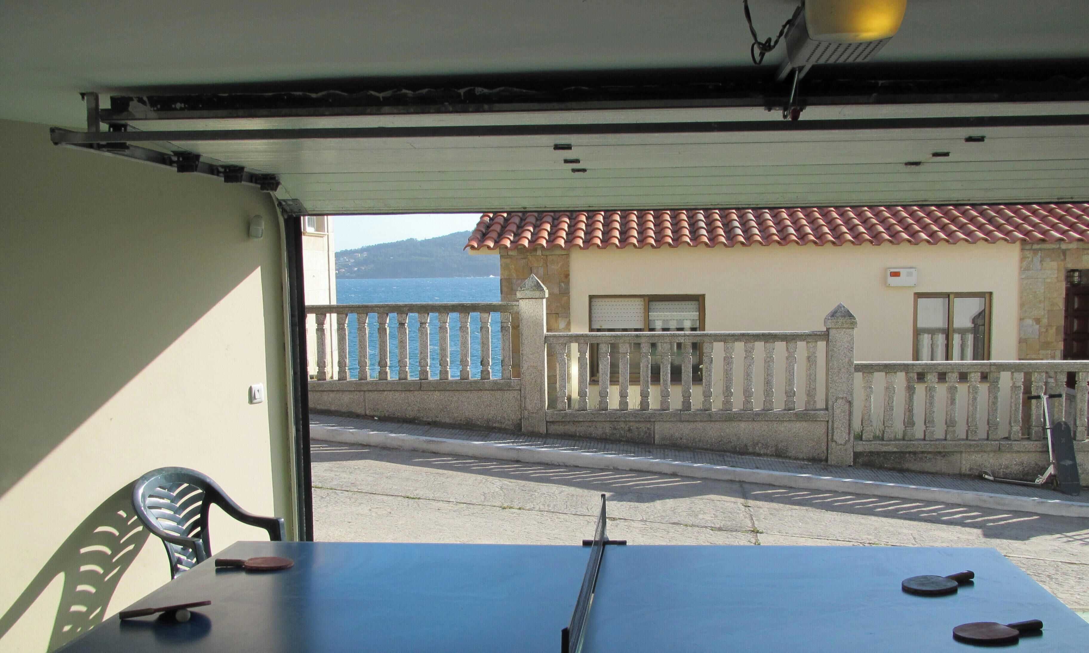 A table tennis table with paddles and a ball, a chair, and a view of a street with a building and a fence.