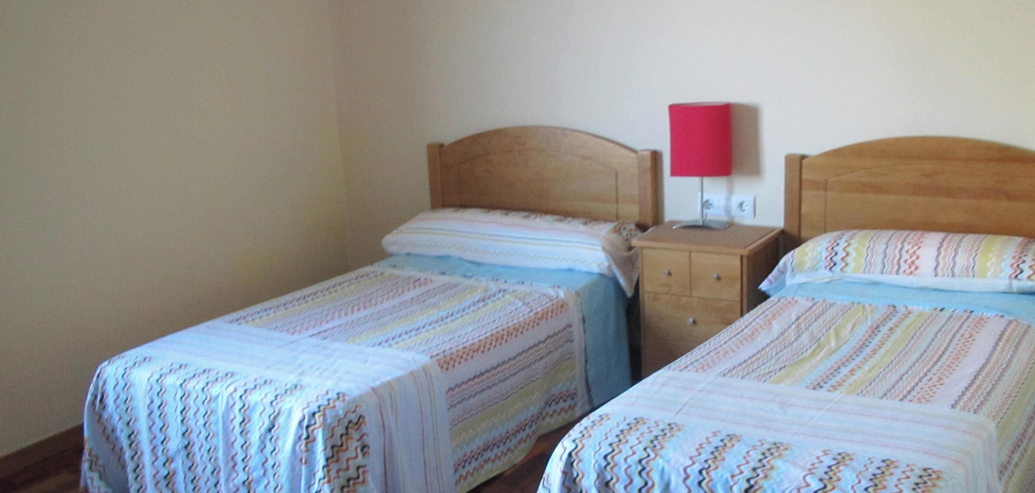 Dos camas individuales con ropa de cama a rayas, una mesita de noche de madera con una lámpara roja y un cabecero de madera.