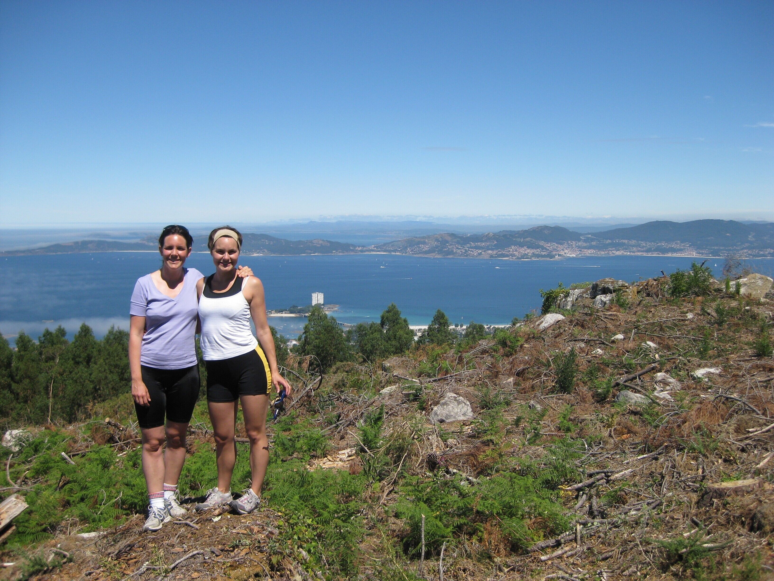 Two people standing on a rocky hillside with a view of a coastal city and the ocean in the background.