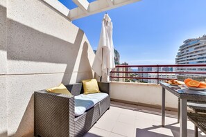En balkon med en fletsofa, et bord med frugt og udsigt til bygninger og havet.