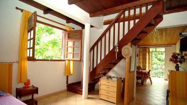 Une pièce spacieuse avec un escalier en bois, une grande fenêtre avec des rideaux, un coin repas avec une table et des chaises, et une vue sur la verdure à l’extérieur.