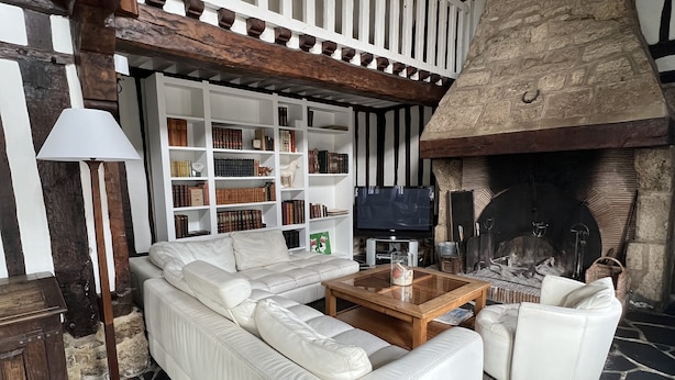 A modern living room with a white sofa, a coffee table, and a fireplace. There is a bookshelf filled with books and a lamp. The room has a stone floor and exposed wooden beams. - Full of charm, authentic and renovated cottage Honfleur (Normandy) (Honfleur)