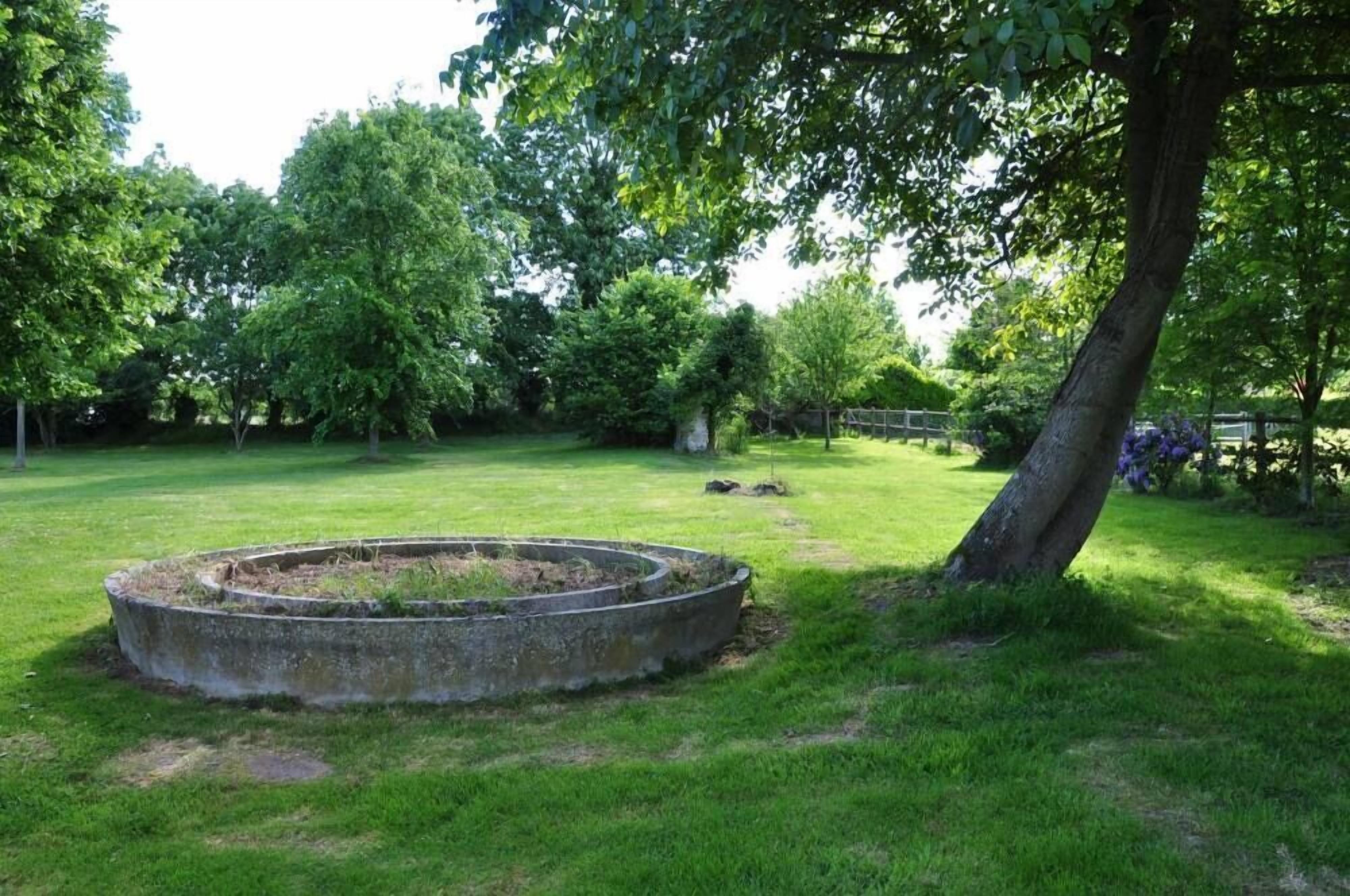 A circular concrete structure in a grassy area with trees and a fence in the background.