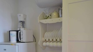 Et kompakt køkken med et hvidt køleskab, en mikroovn og en kaffemaskine på bordpladen. Der er et lille træbord og en hvid reol med kopper og en elkedel.