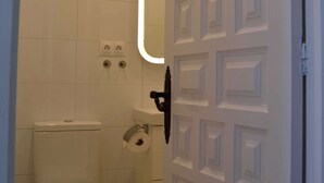 Et badeværelse med toilet, skraldespand, spejl og en dør med et geometrisk mønster.