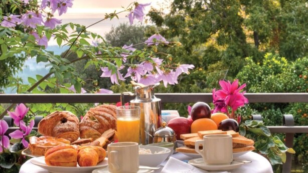 Una tavola per la colazione con pasticcini, frutta e una caraffa di succo d'arancia su un balcone affacciato su alberi e fiori.