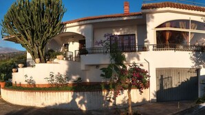Una casa a due piani con un grande cactus nel giardino, un balcone con ringhiera e una montagna sullo sfondo.