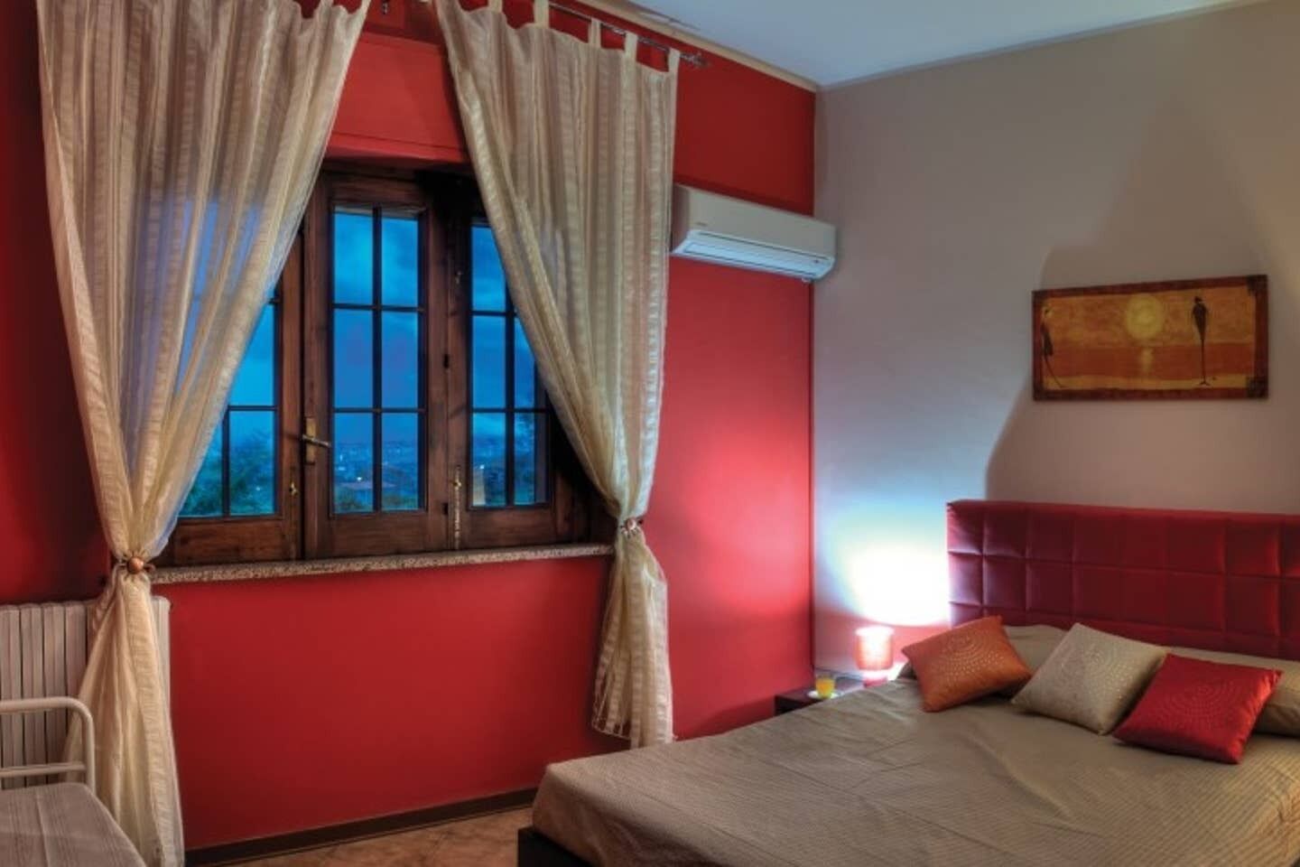A bedroom with a red wall, a window with curtains, a bed with pillows, and a nightstand with a lamp.