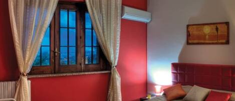 A bedroom with a red wall, a window with curtains, a bed with pillows, and a nightstand with a lamp.