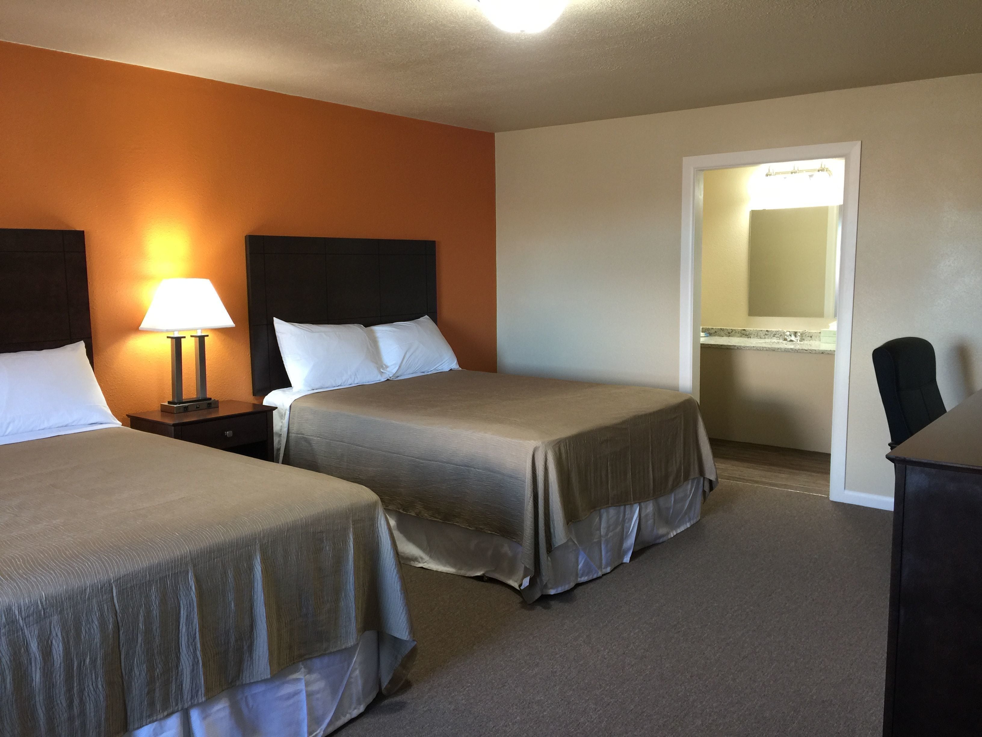 A hotel room with two beds, a nightstand with a lamp, a desk, and a bathroom visible through an open door.