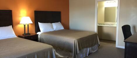 A hotel room with two beds, a nightstand with a lamp, a desk, and a bathroom visible through an open door.