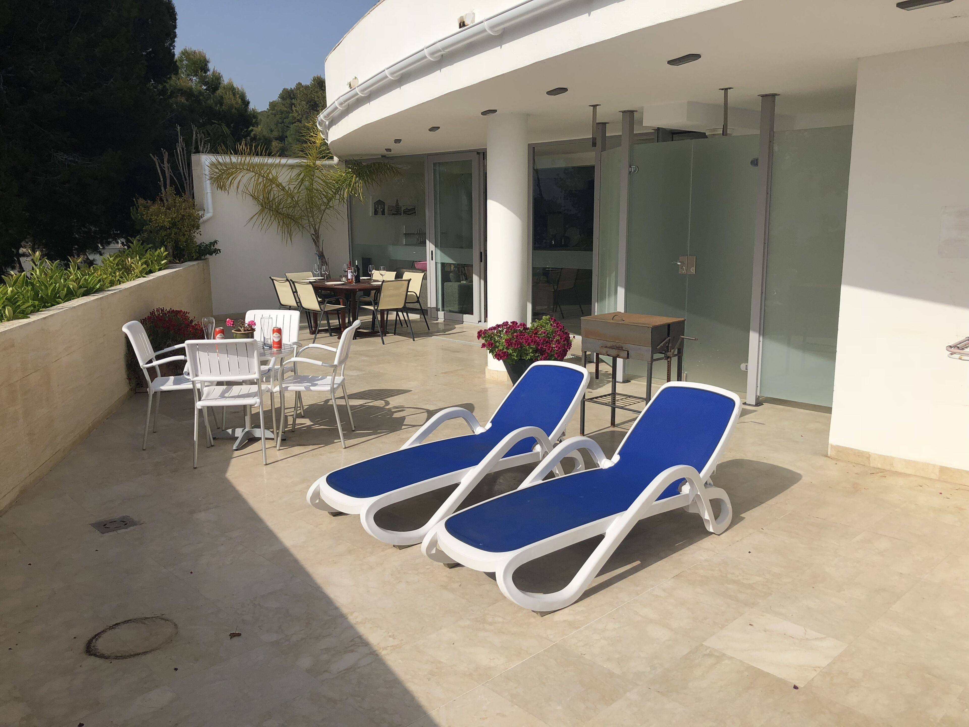 A sunlit outdoor patio with two blue lounge chairs, a table with chairs, and a barbecue grill.