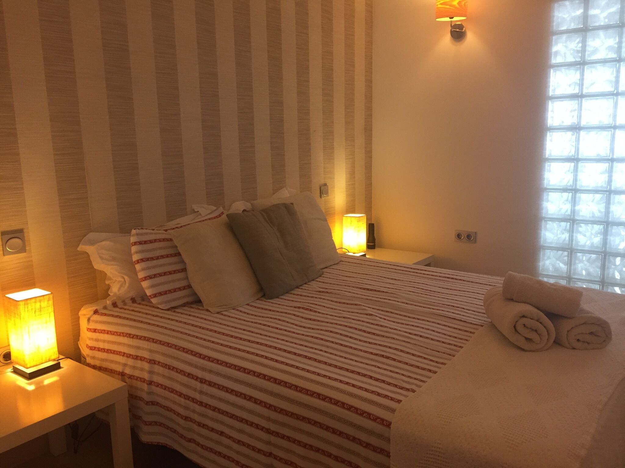 Une chambre d’hôtel avec une couvre-lit à rayures, des oreillers, une serviette enroulée, des lampes de chevet et une fenêtre laissant entrer la lumière naturelle.