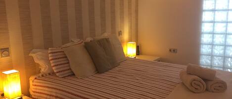 Une chambre d’hôtel avec une couvre-lit à rayures, des oreillers, une serviette enroulée, des lampes de chevet et une fenêtre laissant entrer la lumière naturelle.