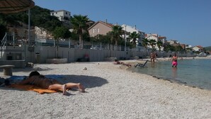 Een kiezelstrand met mensen die zonnebaden en zwemmen, een palmboom en huizen op de achtergrond.