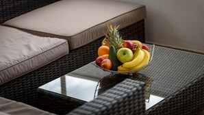 Een rieten bank met beige kussens, een glazen tafel met een fruitschaal met appels, bananen en sinaasappels.