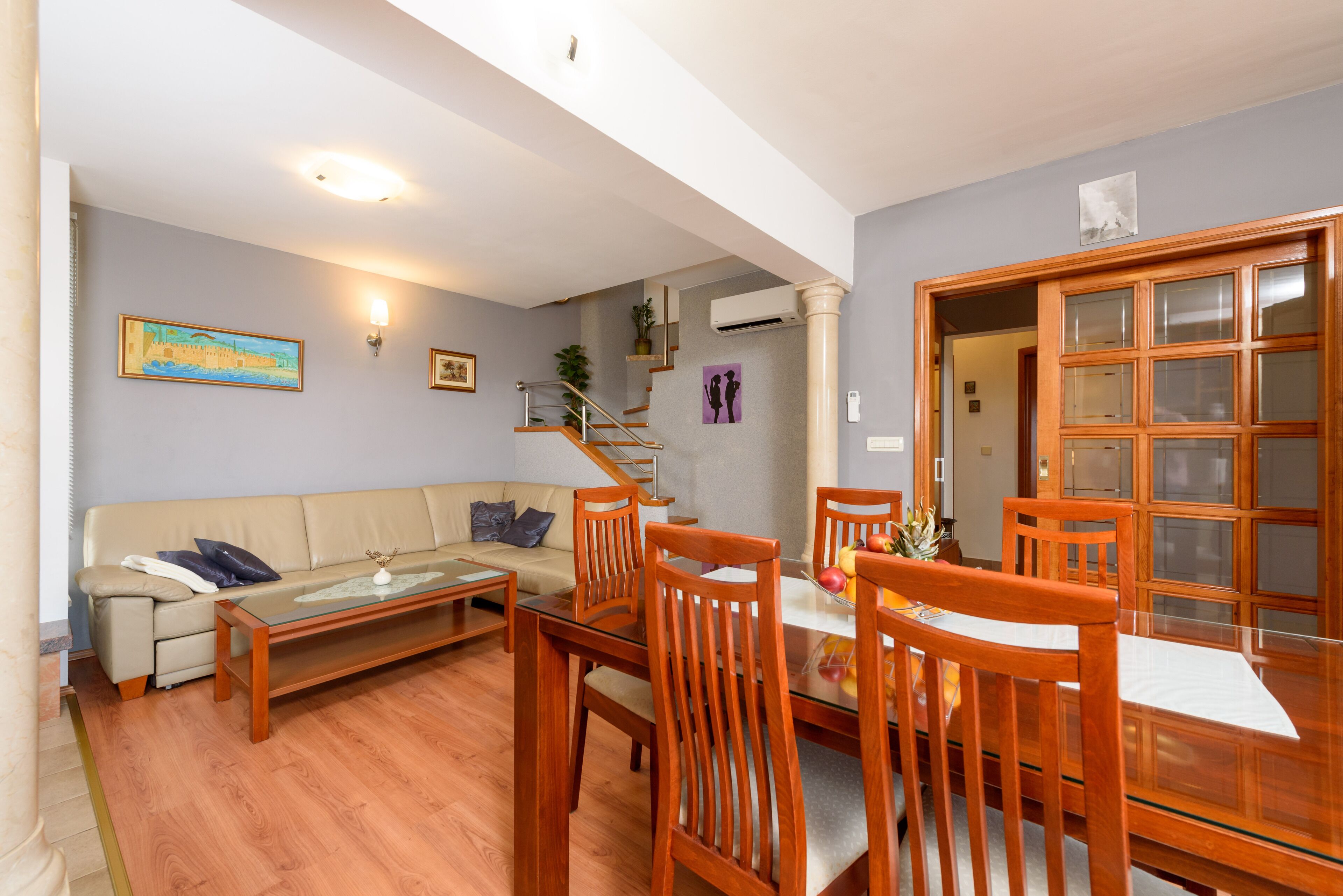 Een woonkamer met een bank, salontafel, houten eettafel en stoelen. Er is een trap die naar een hoger gelegen niveau leidt.