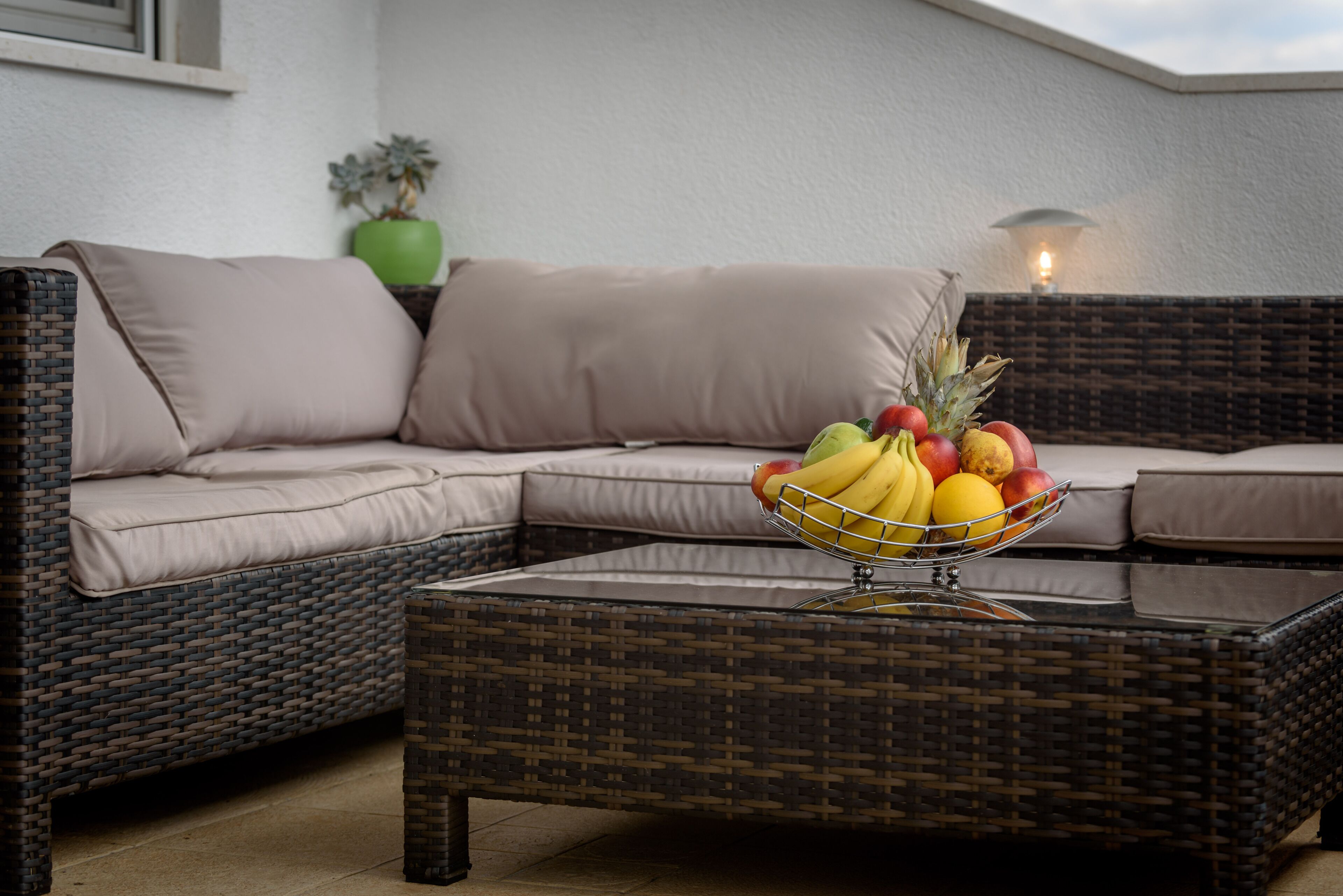 A wicker sofa with a fruit bowl on a matching ottoman, a green plant, and a wall-mounted lamp.