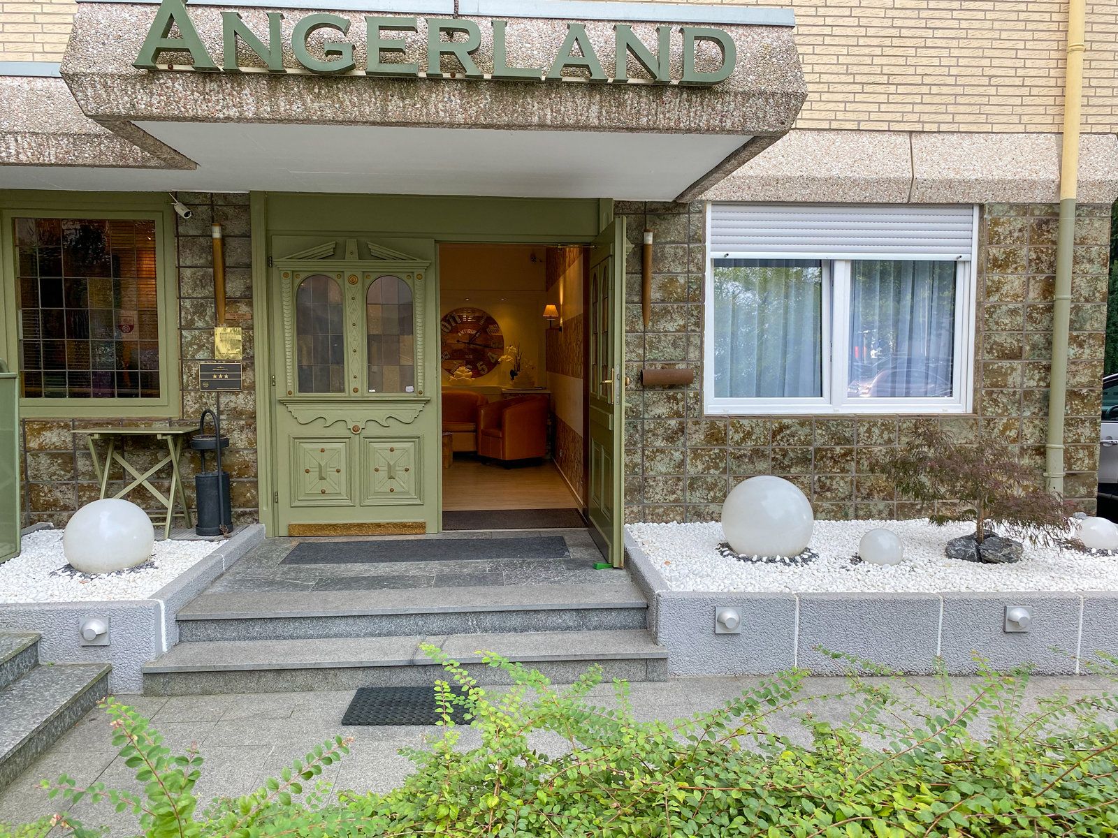 Ein Gebäude mit einem Schild, auf dem „ANGERLAND“ steht, am Eingang. Vor der Tür führen Treppen hinauf, und es gibt eine kleine Gartenanlage mit weißen, kugelförmigen Dekorationen.
