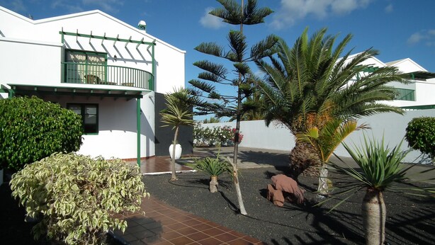 Uma casa moderna com varanda, cercada por diversas plantas e um pátio com piso de azulejos.