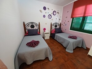 Ein Zimmer mit zwei Betten, jeweils mit grauer Bettdecke und einem Zierkissen. Ein kleiner Nachttisch mit Lampe steht bereit, und die Wände sind mit runden und floralen Stickern verziert.