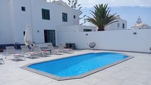 Piscina retangular com deck ladrilhado, rodeada por paredes brancas e uma palmeira.