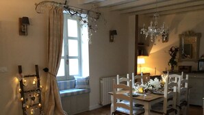 Un coin repas chaleureux avec une table en bois dressée pour quatre personnes, entourée de chaises blanches. Une fenêtre avec des rideaux laisse entrer la lumière naturelle, et un lustre est suspendu au-dessus de la table.