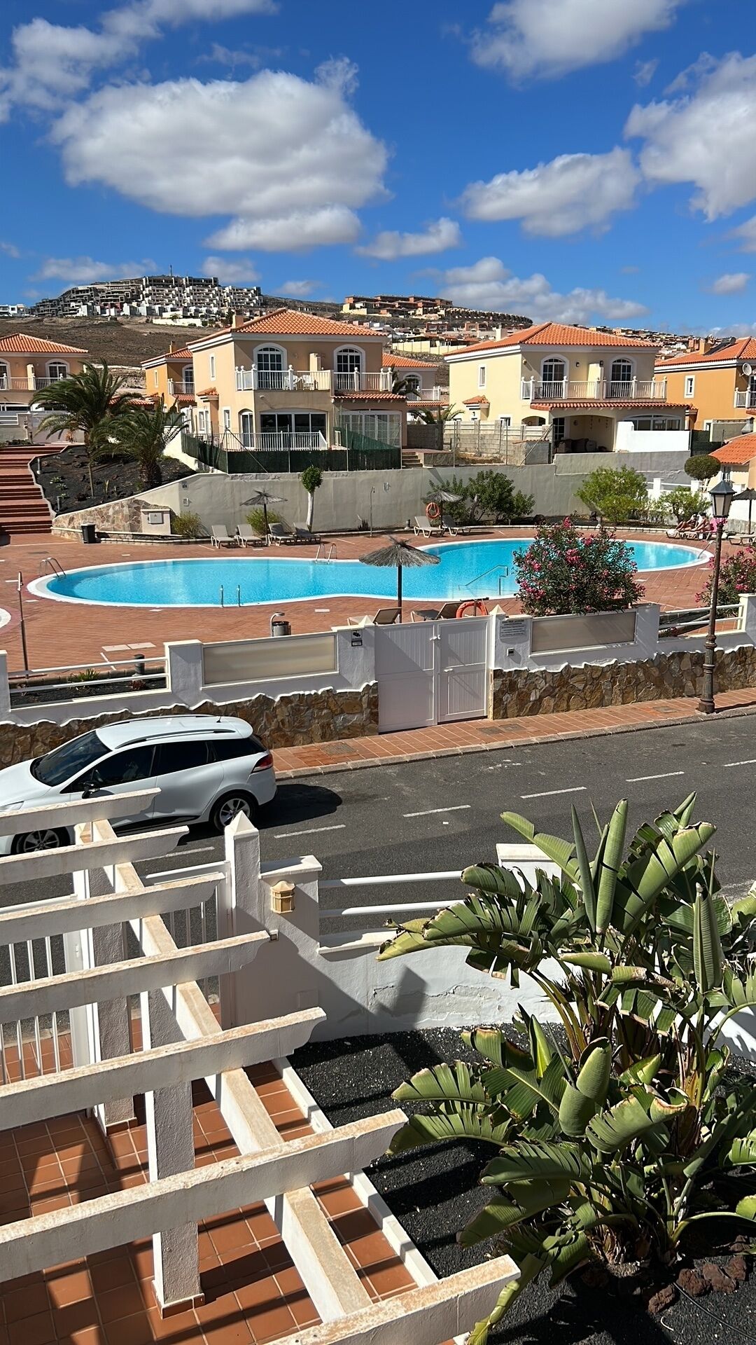 A residential area with a swimming pool, a white car parked in the foreground, and houses with red-tiled roofs in the background.